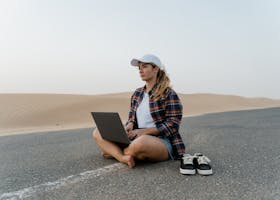 Young woman in plaid shirt using laptop on desert road, embracing digital nomad lifestyle.